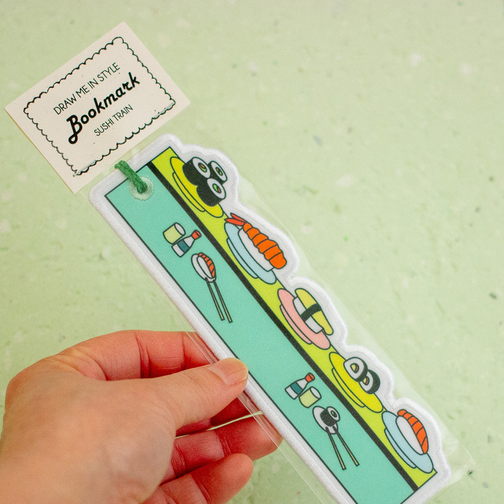 A hand holds a sushi-themed bookmark in its clear plastic packaging against a light green speckled background. The bookmark features cartoon sushi illustrations on a mint green panel.