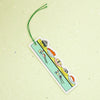 A rectangular fabric bookmark with a green hanging string, featuring cartoon sushi characters on a light green background, set against a textured pale green surface.
