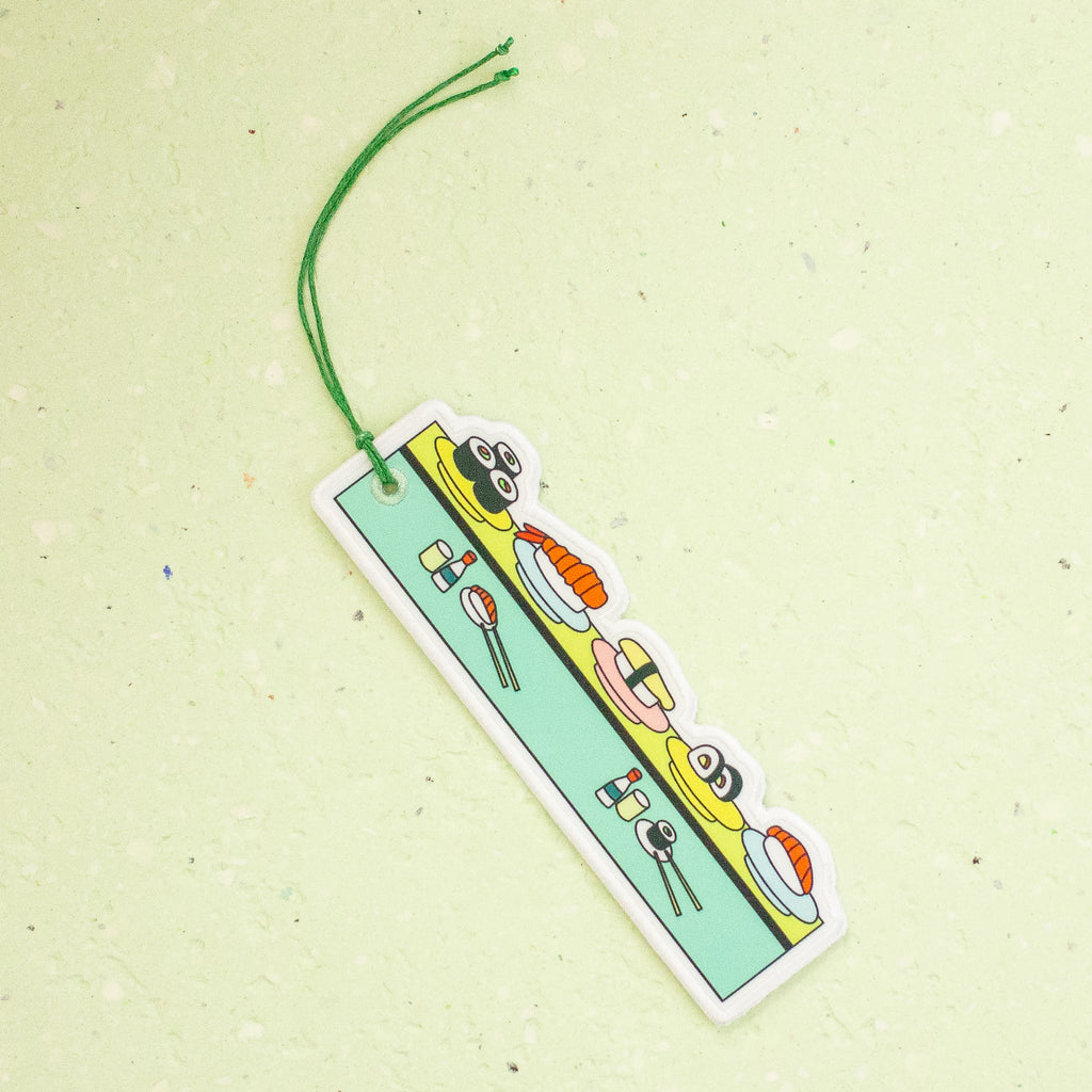 A rectangular fabric bookmark with a green hanging string, featuring cartoon sushi characters on a light green background, set against a textured pale green surface.