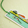 A close-up of a colourful sushi-themed acrylic keychain or tag with a green hanging string, featuring illustrations of sushi rolls, a shrimp roll, chopsticks, and condiments on a light green textured background.
