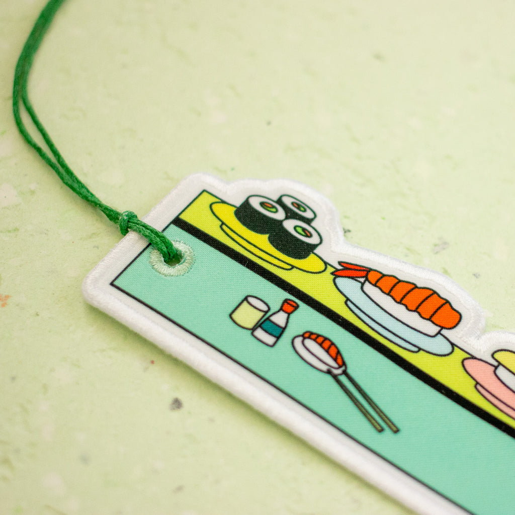A close-up of a colourful sushi-themed acrylic keychain or tag with a green hanging string, featuring illustrations of sushi rolls, a shrimp roll, chopsticks, and condiments on a light green textured background.