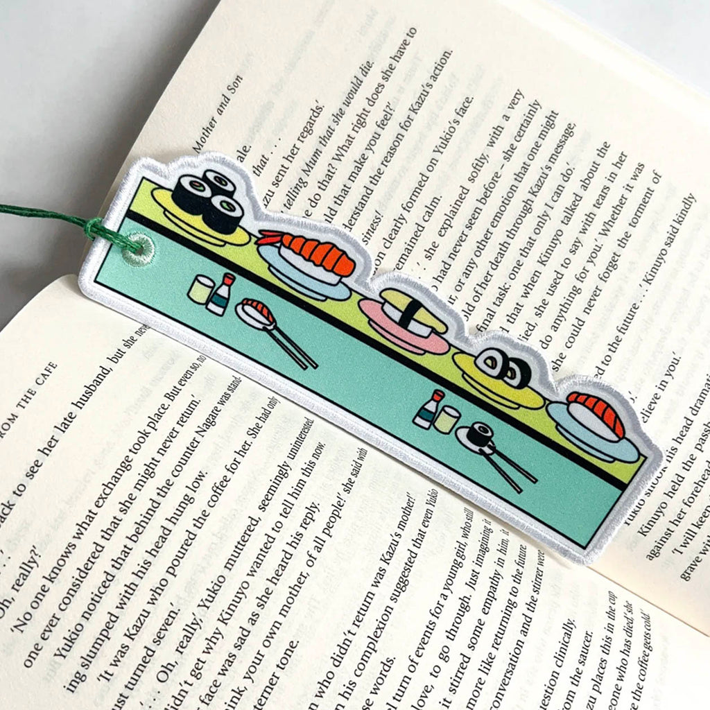 A sushi-themed bookmark with a cartoon sushi illustration and a green string, shown resting on an open book.