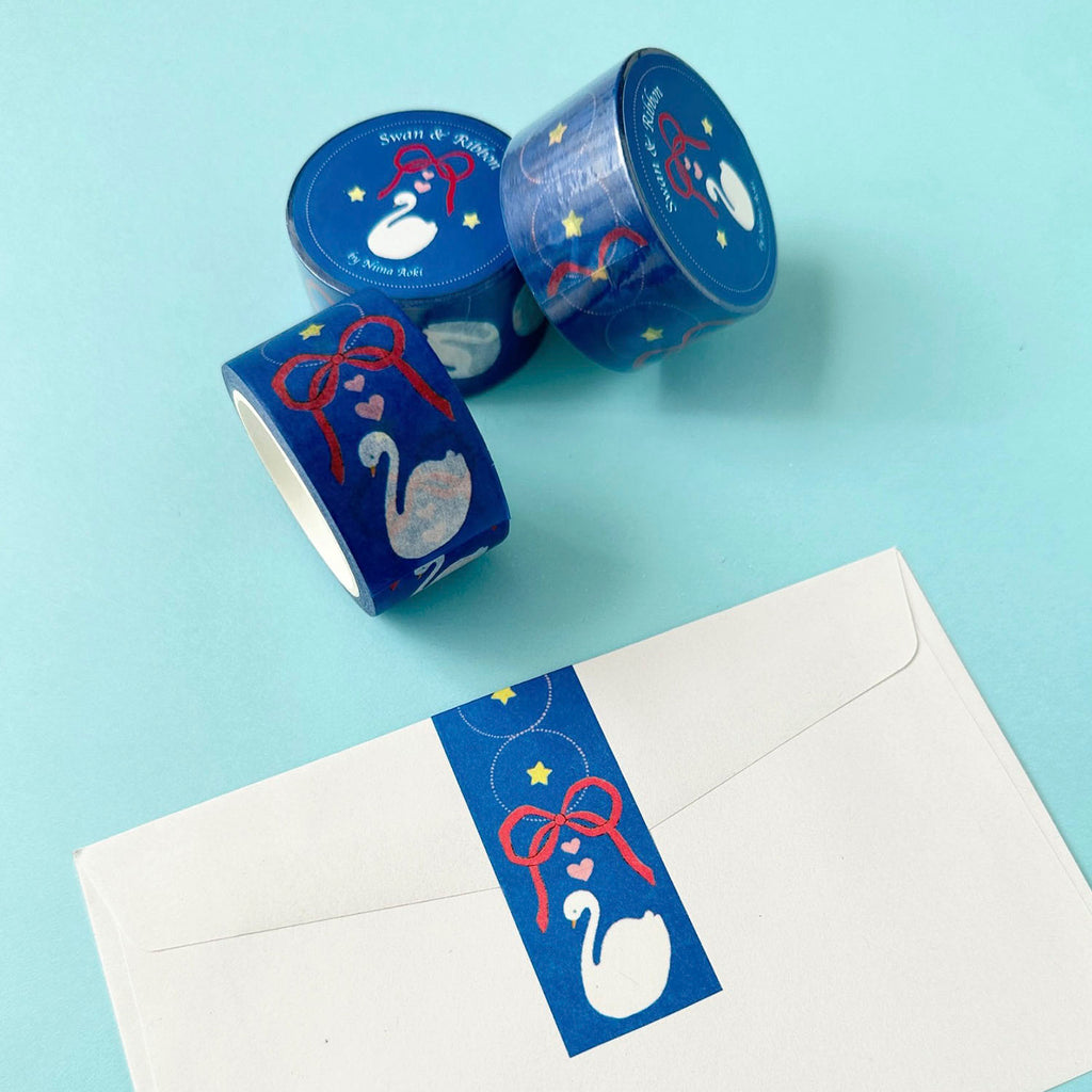 Three rolls of decorative washi tape with a blue, red, and white swan design are arranged on a light blue background next to a white envelope featuring a matching blue label.