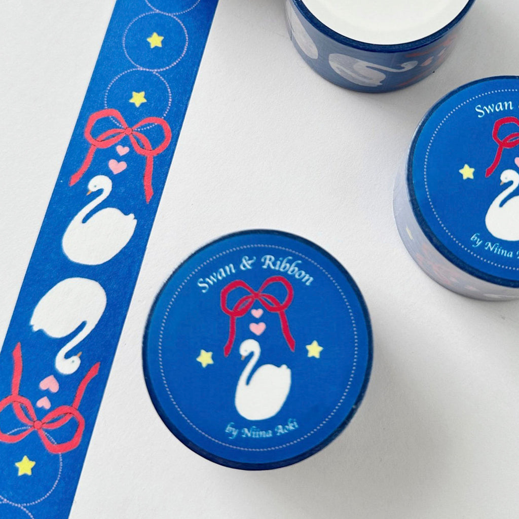 Blue decorative tape with a repeating pattern of white swans, red bows, and yellow stars, shown unrolled alongside two matching rolls on a white background. 