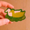 A hand holding a die-cut sticker featuring a yellow duck swimming in green water, with a white duck next to it. The background is a warm, solid tan surface.