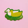 A die-cut sticker featuring ducks swimming in green water, set against a solid pink background.