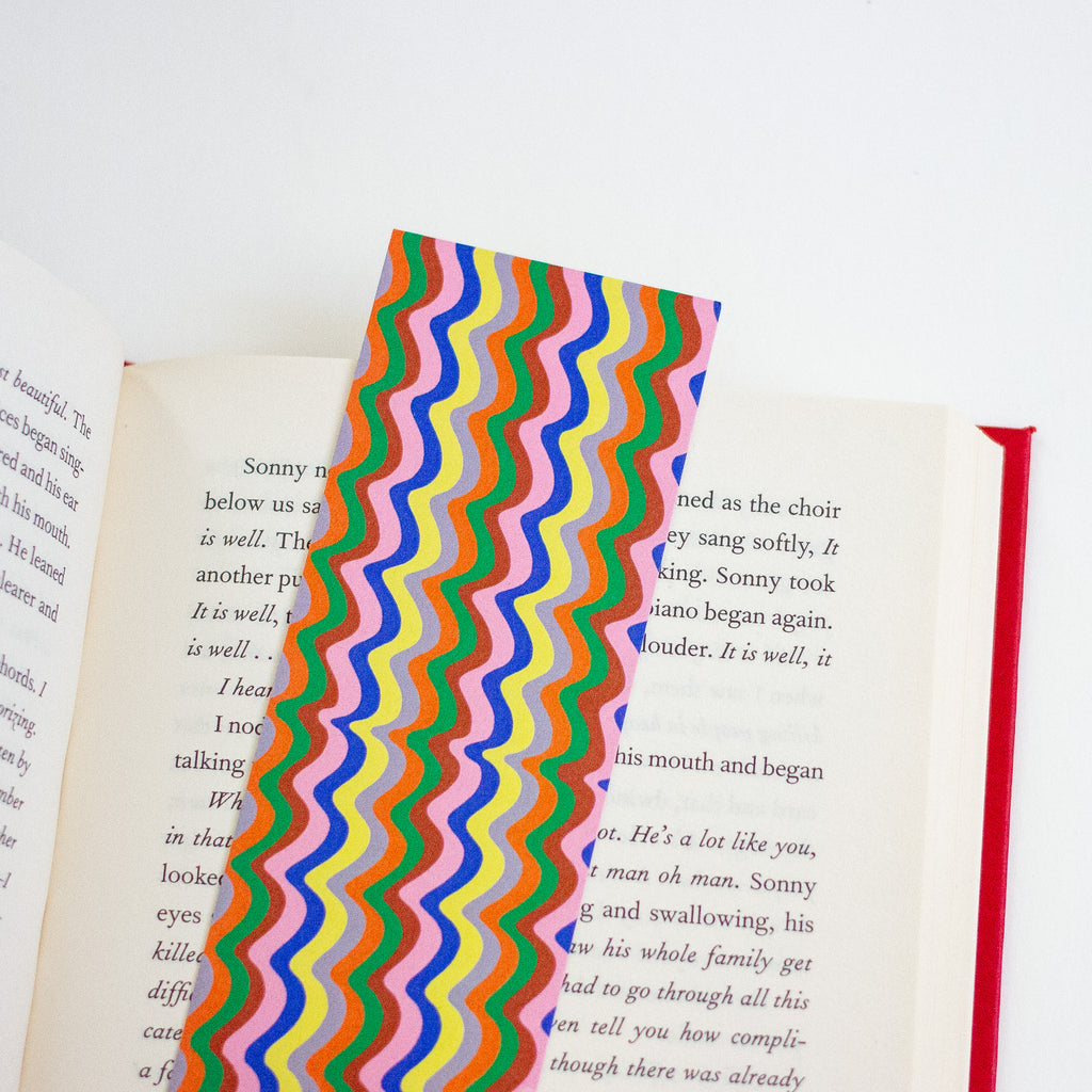 A colourful wavy stripe pattern bookmark with pink, orange, brown, yellow, grey, green, and blue,  shown resting on an open book against a plain white background.