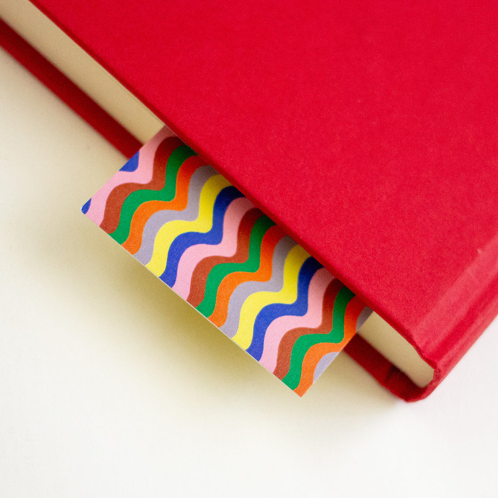 A colourful wavy stripe pattern bookmark with pink, orange, brown, yellow, grey, green, and blue, shown peeking out of a closed book against a plain white background.