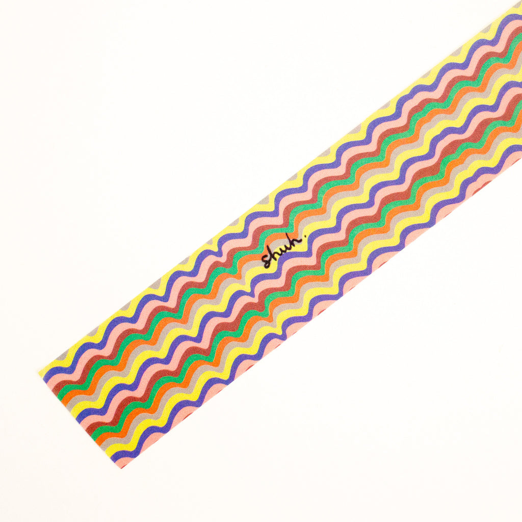 A diagonal strip of colourful wavy-patterned tape on a plain white background, featuring the brand name 'shuh.'