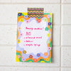 A colourful handwritten note tapped to a white speckled tile wall. The note features a whimsical wavy border decorated with small flowers in multiple colours, with handwritten pink text reading 'Yummy cookies! Get: almond meal, tahini, maple syrup'.