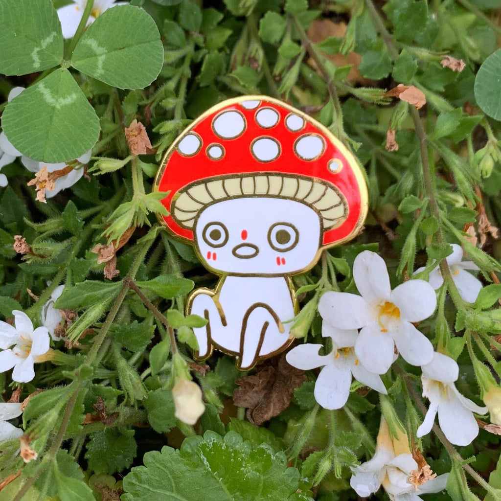 Enamel pin of a mushroom character surrounded by green leaves and white flowers