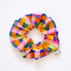 Fabric scrunchie with a bright and colourful tartan-inspired pattern in shades of orange, pink, green, purple, yellow, and blue, displayed in a circular shape on a white background.