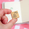 A hand holds a small wooden rubber stamp featuring a black ink design of a cup of coffee and a cupcake. The background is a soft-focus cream-colored notebook with pink accents.