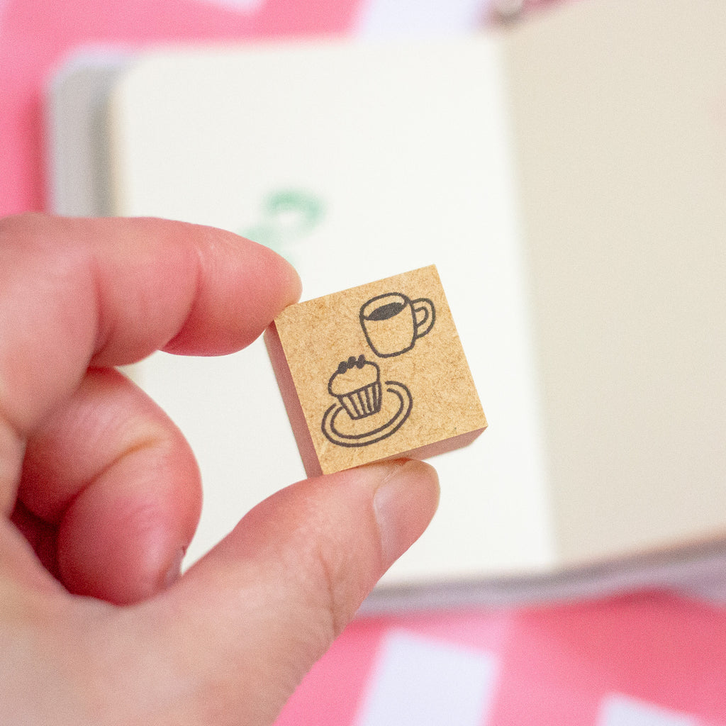 A hand holds a small wooden rubber stamp featuring a black ink design of a cup of coffee and a cupcake. The background is a soft-focus cream-colored notebook with pink accents.
