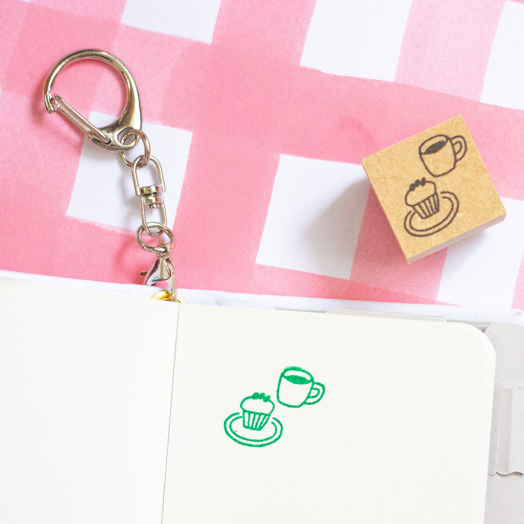 A small wooden rubber stamp with a cup and muffin design, shown next to a metal clasp and keychain, with the design stamped green on a white notebook page against a pink and white checkered background.