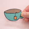 A hand holding a teacup shaped ceramic brooch with a brown interior and an orange tea bag, against a solid pale pink background.