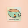 A hand illustrated light teal mug with a brown interior, a yellow-orange tea bag on a square ceramic tile card with the text 'juliebceramics.com.au'