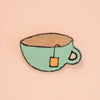 A hand-drawn style brooch shaped like a light green teacup with a brown interior, black outlines, and a small orange tea bag tag, shown on a solid pale pink background.