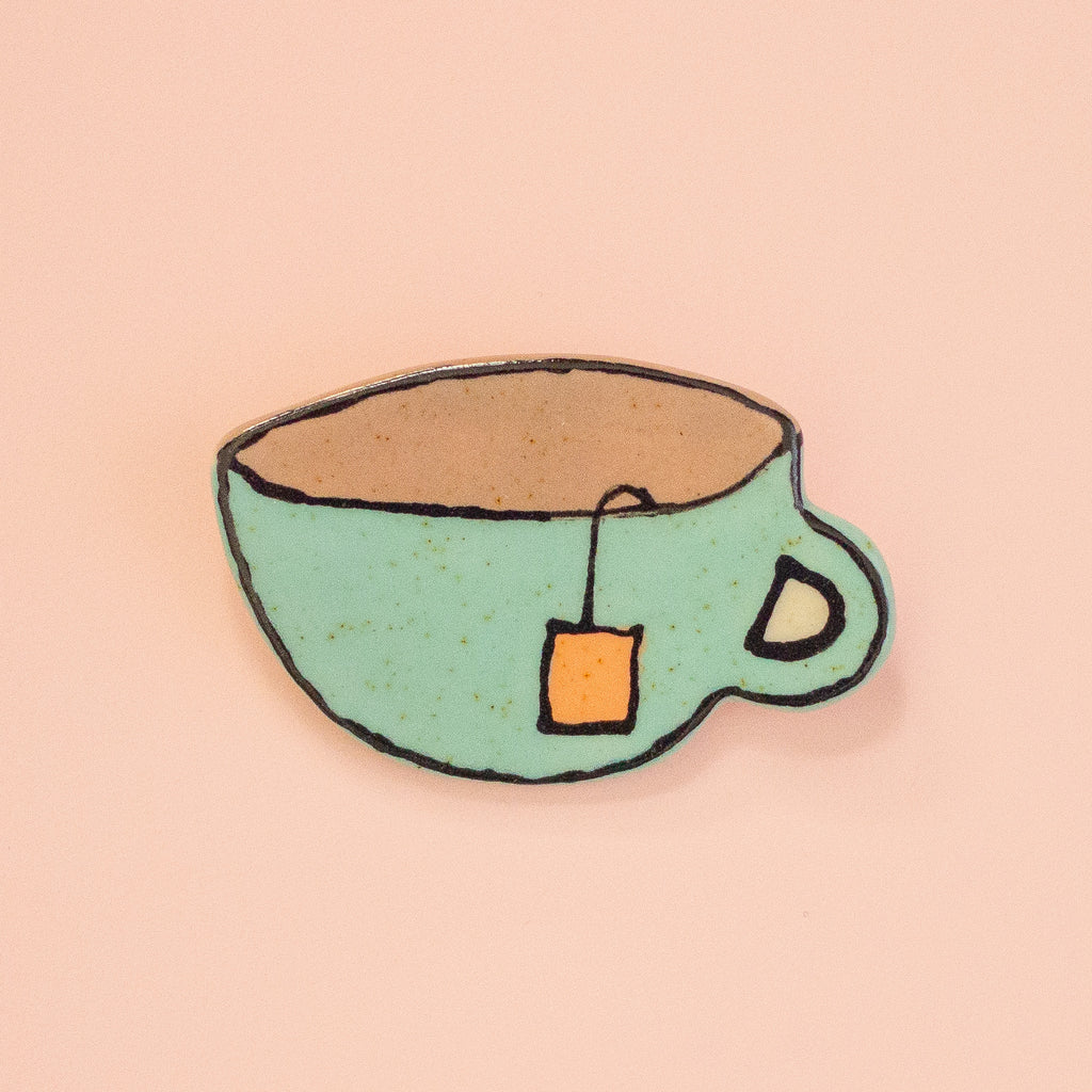 A hand-drawn style brooch shaped like a light green teacup with a brown interior, black outlines, and a small orange tea bag tag, shown on a solid pale pink background.