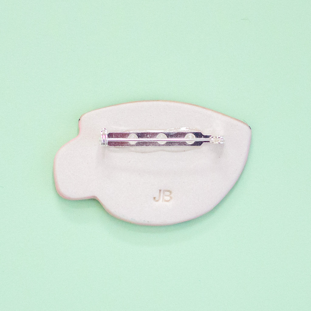 A brooch with a silver metal pin bar and the letters 'JB' embossed on the front, shown on a mint green background.
