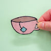 A hand holding a hand-drawn pink teacup illustration with black outlines, a brown interior, and a light blue tea bag tag, against a pale green background.