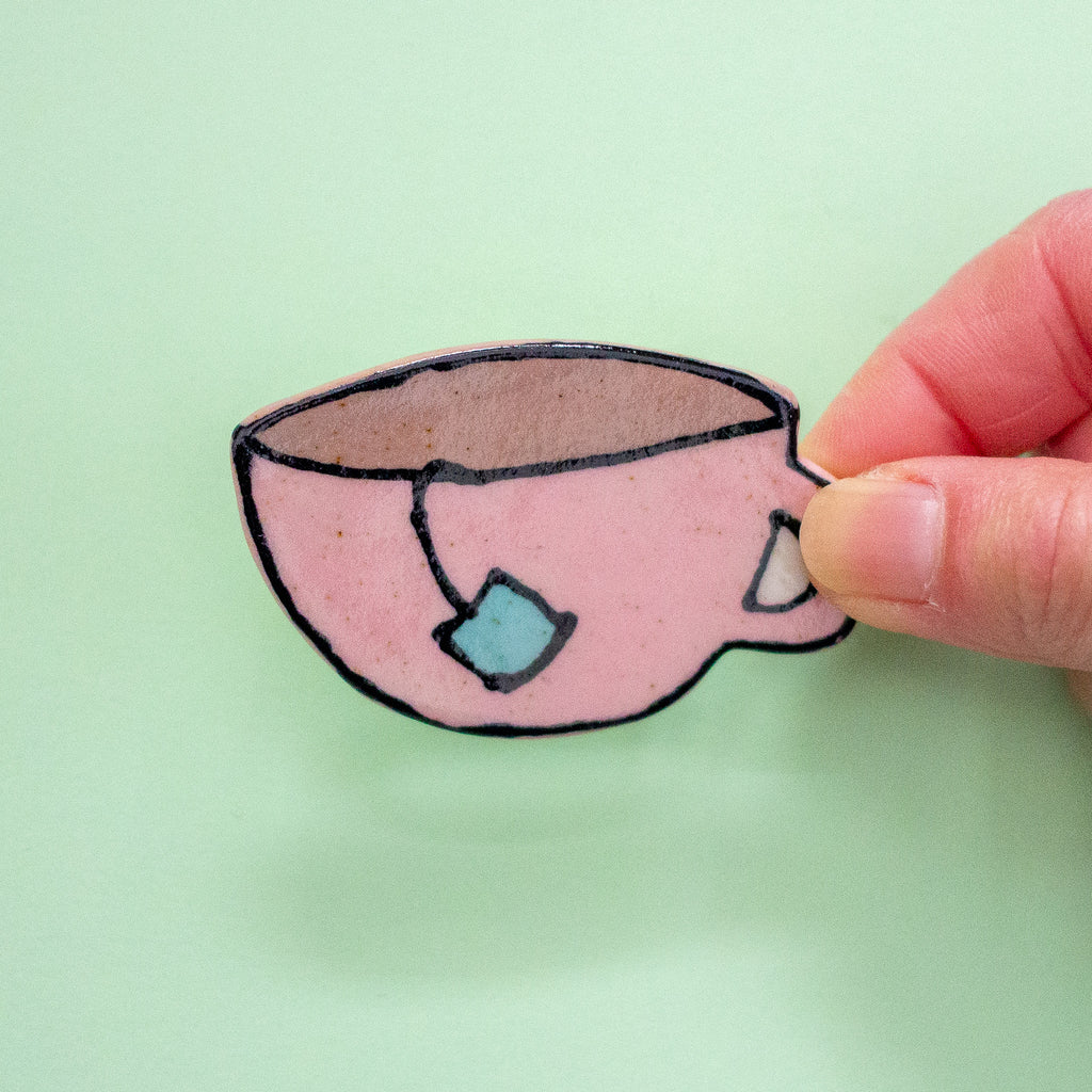 A hand holding a hand-drawn pink teacup illustration with black outlines, a brown interior, and a light blue tea bag tag, against a pale green background.
