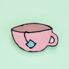 A pink ceramic teacup brooch with black outlines, a brown interior, and a light blue tea bag tag, shown on a mint green background.