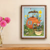 Framed artwork of a teapot with garden scene on a wall above a wooden surface with flowers.