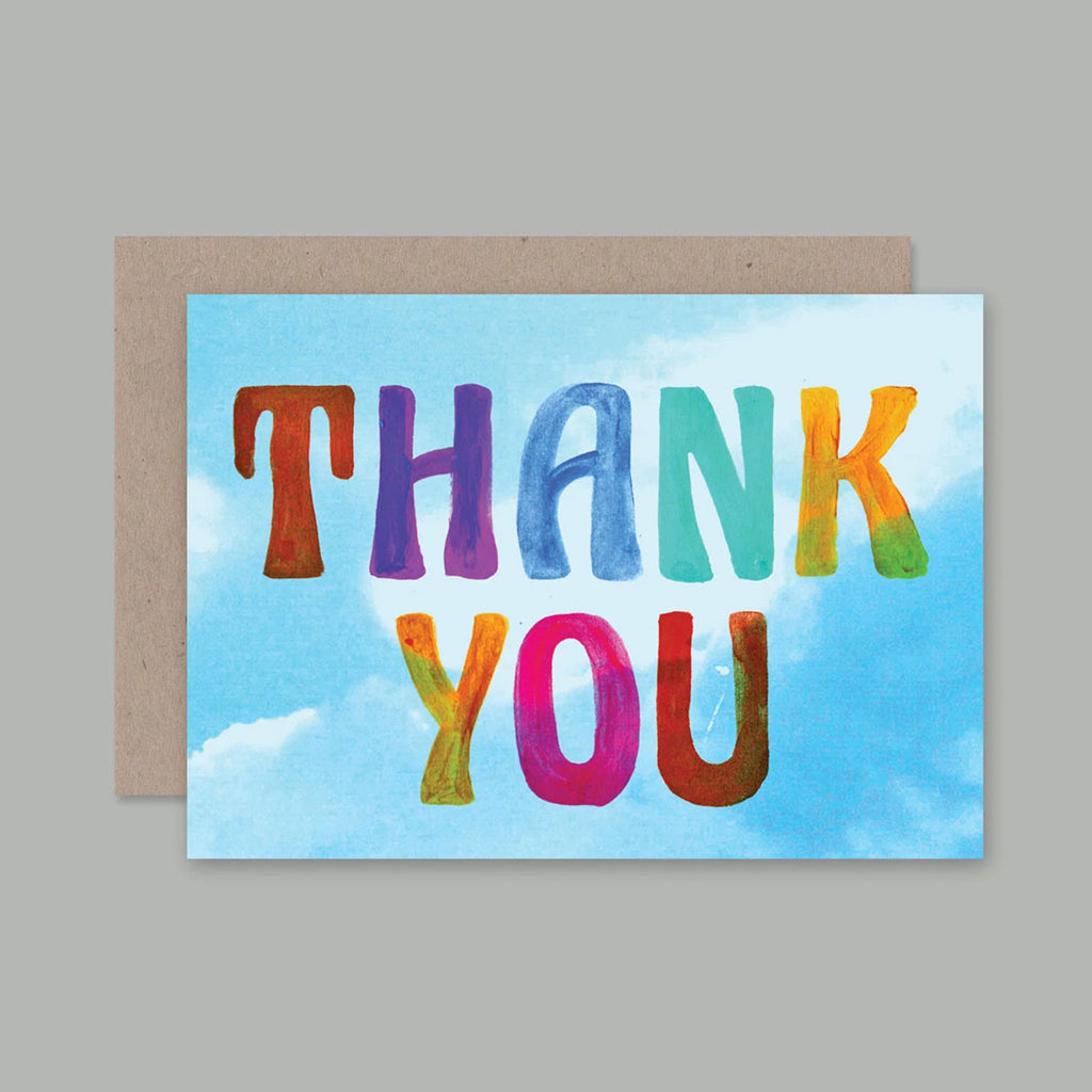 A greeting card with the words “THANK YOU” in large, colouful hand-painted style letters over a light blue sky background with white clouds. A brown kraft envelope is placed behind the card, which is photographed on a solid grey background.