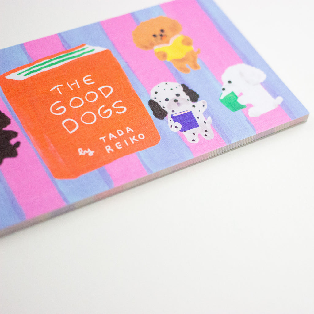 A rectangular notepad featuring a colourful illustration of several dogs reading books on pink and blue vertical stripes, with a central orange book reading 'THE GOOD DOGS' and 'by TADA REIKO'.