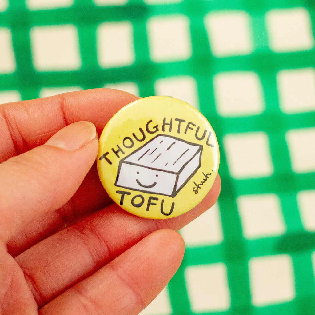 A hand holding a round yellow pinback button with a hand-drawn smiling tofu graphic and the words 'THOUGHTFUL TOFU'. The background is a blurred green and white checkered pattern.