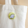 A round pin-back button badge with a light yellow background and a cartoon tofu block illustration, featuring the text 'THOUGHTFUL TOFU' and a small signature, shown pinned to a white canvas tote bag.
