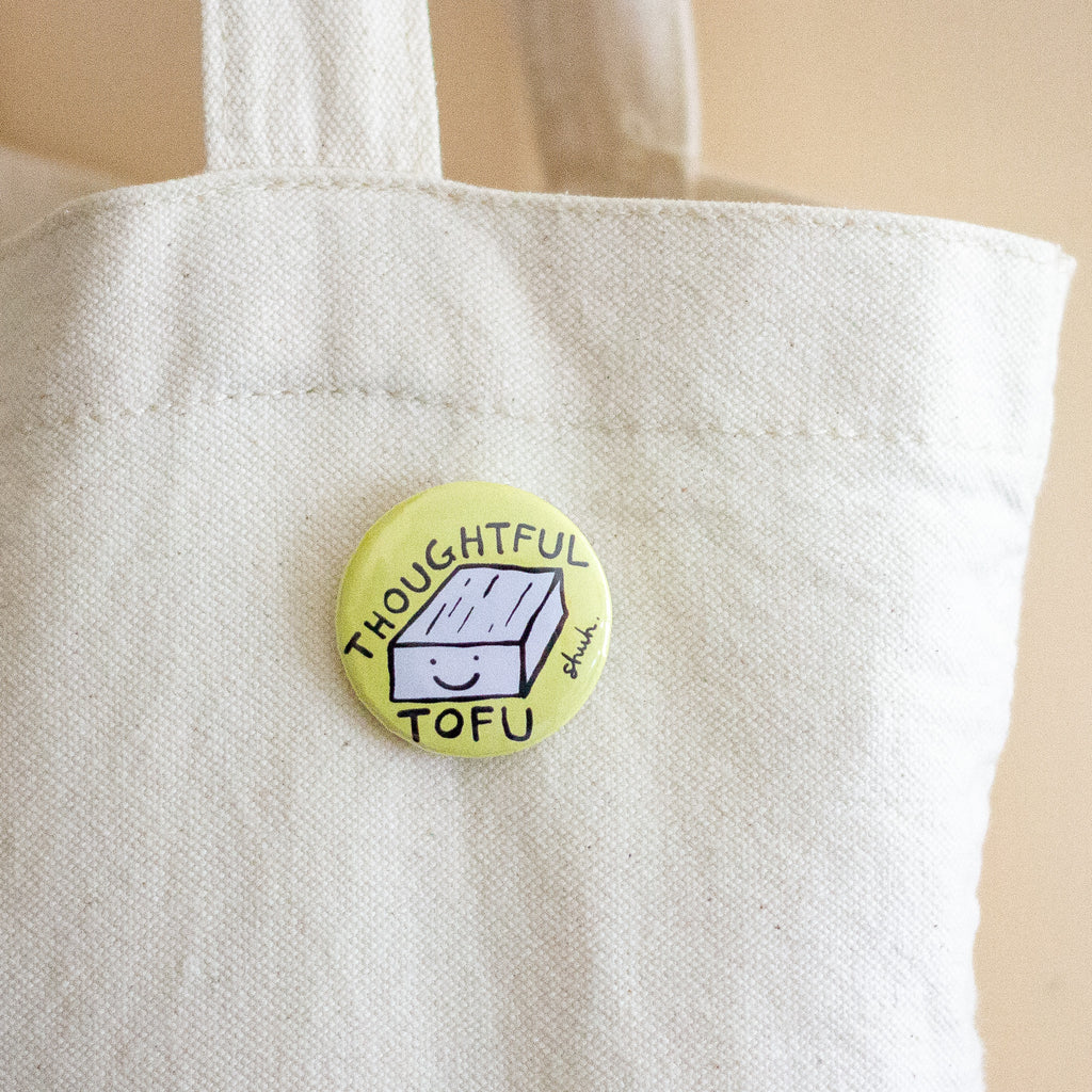 A round pin-back button badge with a light yellow background and a cartoon tofu block illustration, featuring the text 'THOUGHTFUL TOFU' and a small signature, shown pinned to a white canvas tote bag.