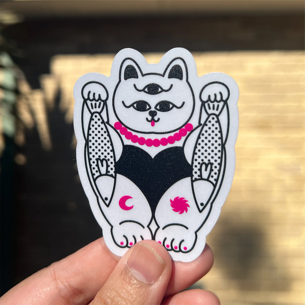 A hand holding a die-cut sticker of a white and black cat with pink accents, set against a blurred outdoor background.