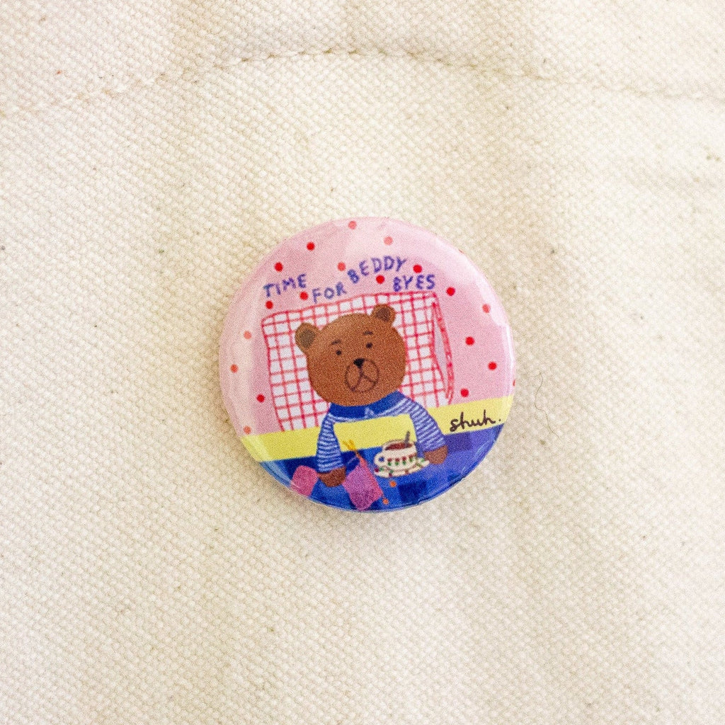 A round pin badge featuring a cartoon teddy bear sitting on a bed with the text 'TIME FOR BEDDY BYES' and a small 'shuh.' logo, pinned onto an off-white canvas tote bag.
