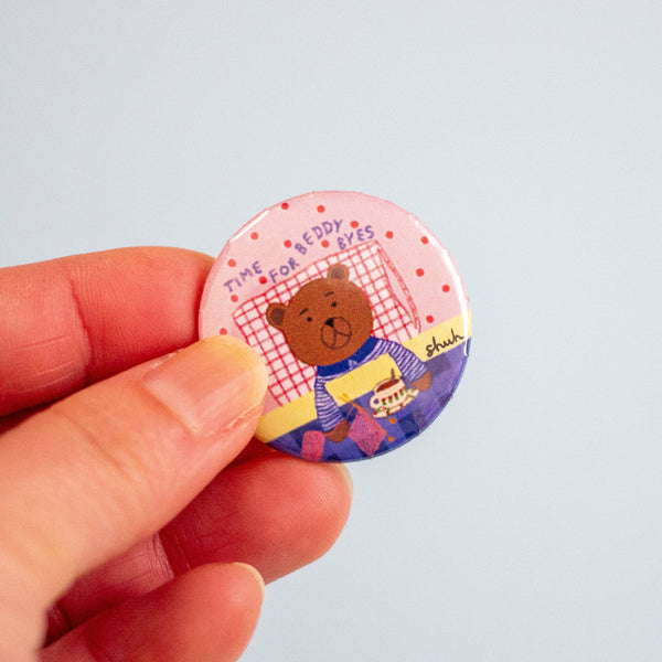 A hand holding a round pin-back button badge featuring a cartoon teddy bear wearing a blue striped sweater, with the text 'TIME FOR BEDDY BYES' on a pink polka-dot background.