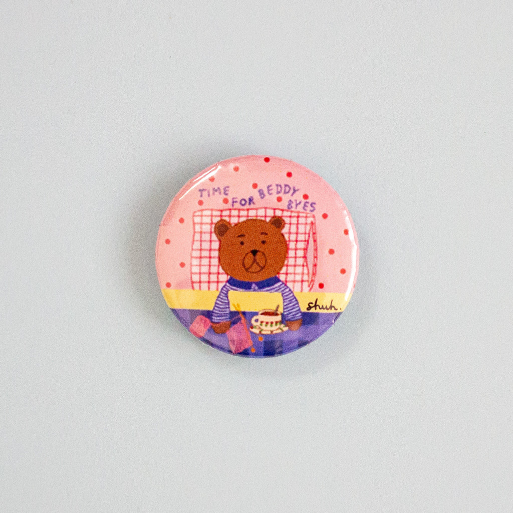A round pinback button featuring a cartoon teddy bear sitting on a bed with the text 'TIME FOR BEDDY BYES' and a pink polka-dot background, shown on a plain light blue surface.