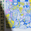 A hand-drawn colourful art print featuring cute cartoon characters, a retro gadgets, and handwritten Japanese text, photographed on a patterned fabric surface.