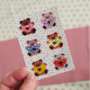 A hand holds a small white sticker sheet featuring six pixel-art teddy bears holding heart shapes, with a pink and cream patterned background.