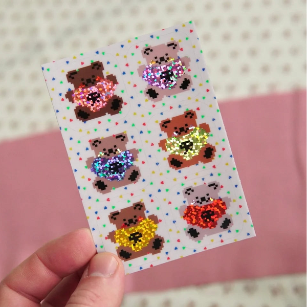 A hand holds a small white sticker sheet featuring six pixel-art teddy bears holding heart shapes, with a pink and cream patterned background.