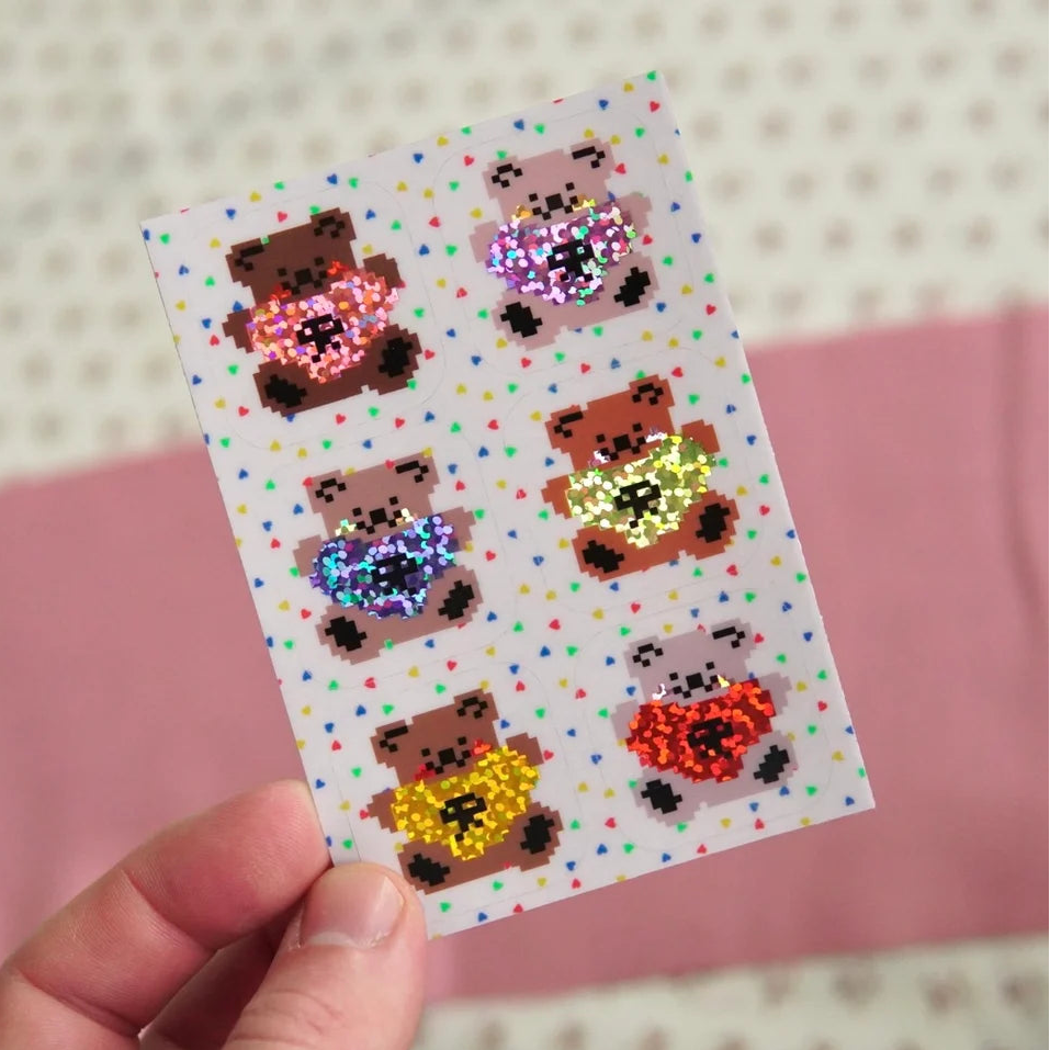 A hand holds a small white sticker sheet featuring six pixel-art teddy bears holding heart shapes, with a pink and cream patterned background.