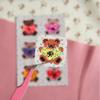 A small square sticker or patch featuring a pixelated brown teddy bear with a glittery gold heart shape, held by pink tweezers in front of a blurred sheet of similar decorative stickers on a pink and cream background.