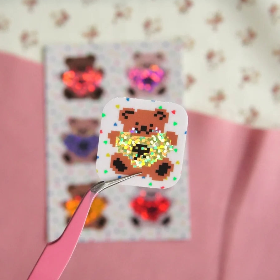 A small square sticker or patch featuring a pixelated brown teddy bear with a glittery gold heart shape, held by pink tweezers in front of a blurred sheet of similar decorative stickers on a pink and cream background.