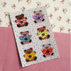 A rectangular sticker sheet featuring six pixelated teddy bears holding colourful glittery heart shapes, displayed diagonally on a pink textured surface over a floral cream background.