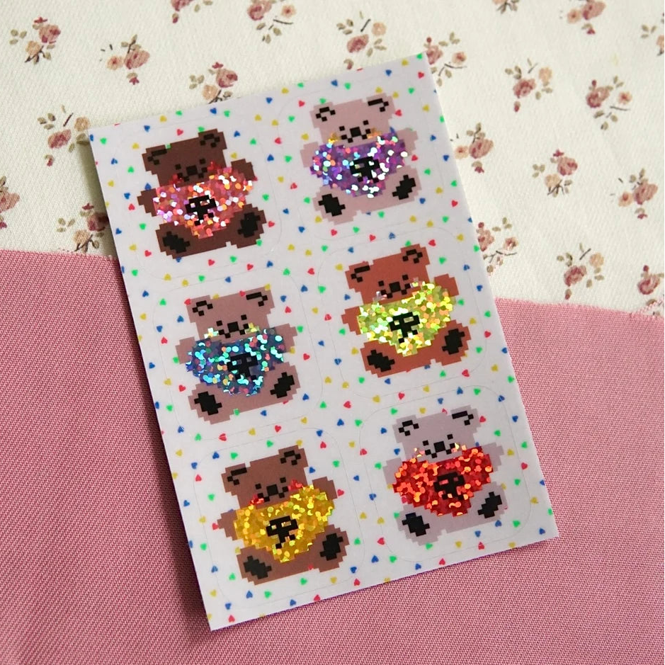 A rectangular sticker sheet featuring six pixelated teddy bears holding colourful glittery heart shapes, displayed diagonally on a pink textured surface over a floral cream background.