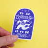 A hand holds a die-cut sticker with a blue-purple and white design that reads 'TO BE WONKY IS TO BE FREE' with a whimsical cartoon character, against a solid yellow background.