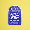 A die-cut sticker featuring a white cartoon character and the text 'TO BE WONKY IS TO BE FREE' on a dark blue-purple arched background, placed on a solid yellow surface.