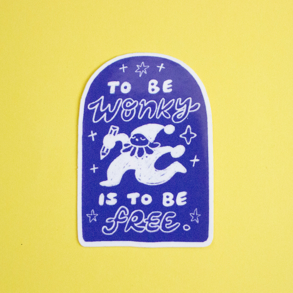A die-cut sticker featuring a white cartoon character and the text 'TO BE WONKY IS TO BE FREE' on a dark blue-purple arched background, placed on a solid yellow surface.