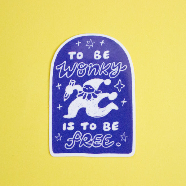 A die-cut sticker featuring a white cartoon character and the text 'TO BE WONKY IS TO BE FREE' on a dark blue-purple arched background, placed on a solid yellow surface.