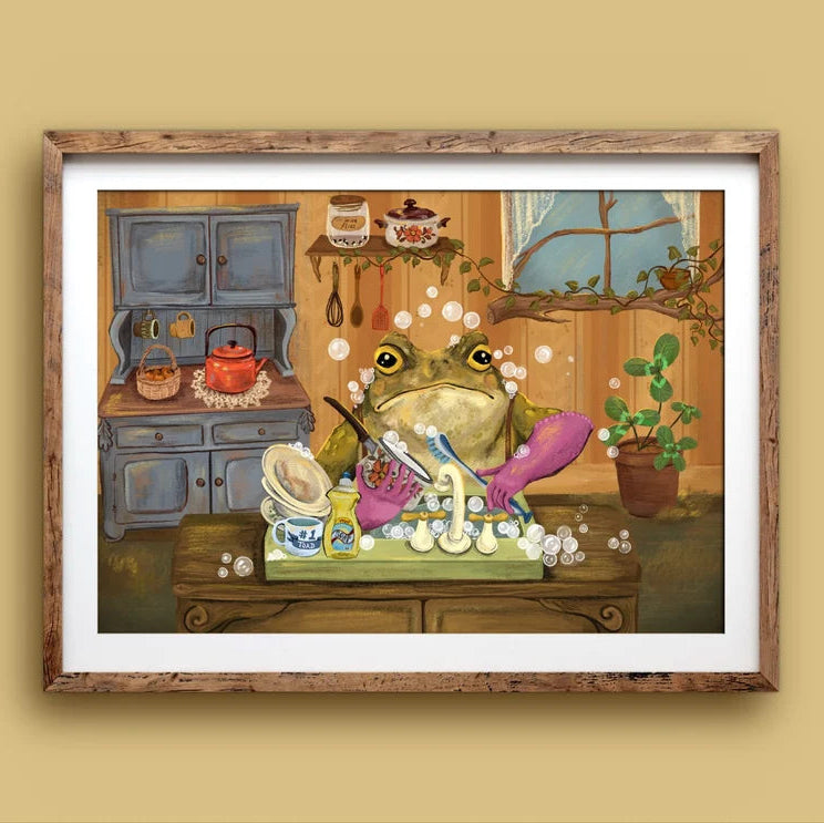 A framed art print featuring a whimsical illustration of a green frog washing dishes in a kitchen setting, mounted on a beige wall.